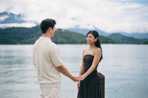 Couple holding hands on a dock by a serene lake with lush green mountains in the background.