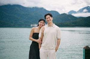 A couple smiles together on a lakeside dock with misty mountains and turquoise water in the background.