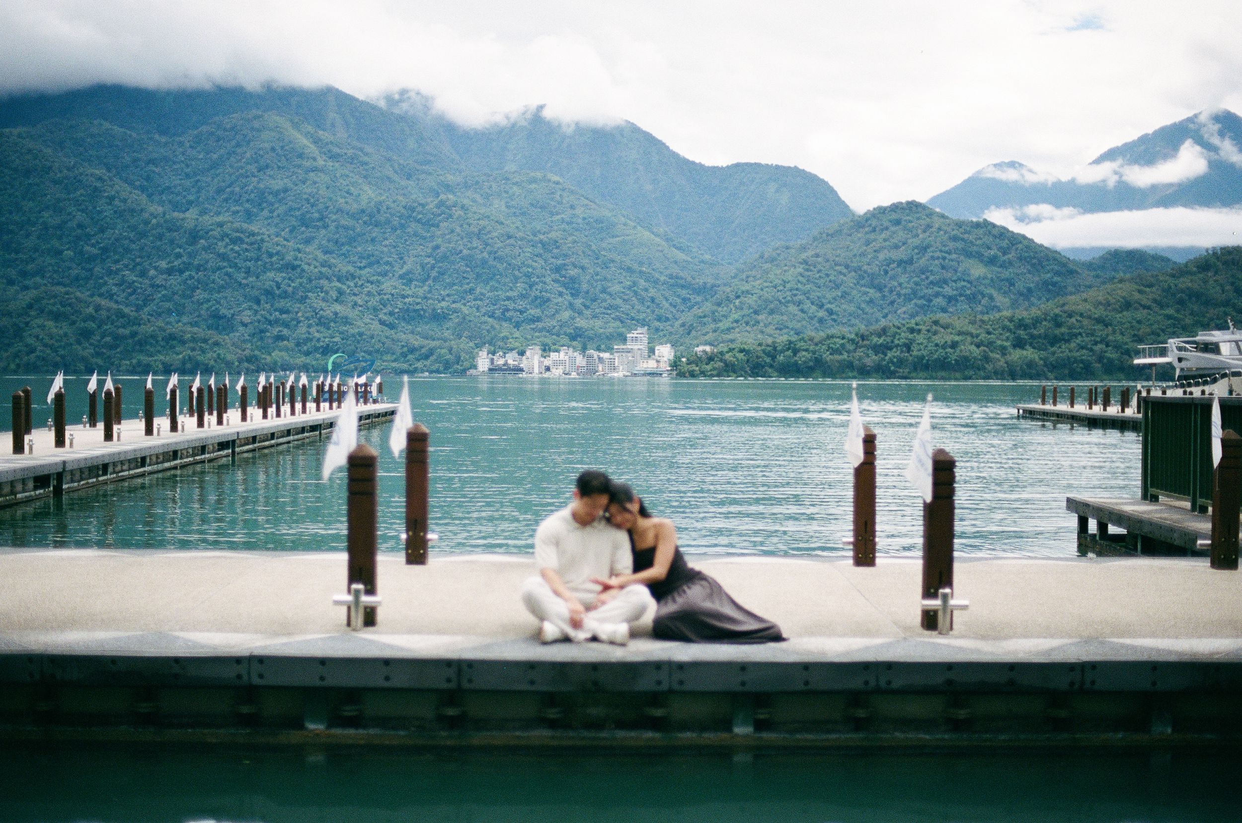 Couple sitting on a wooden dock at Sun Moon Lake, Taiwan, with lush green mountains and calm blue water in the background.