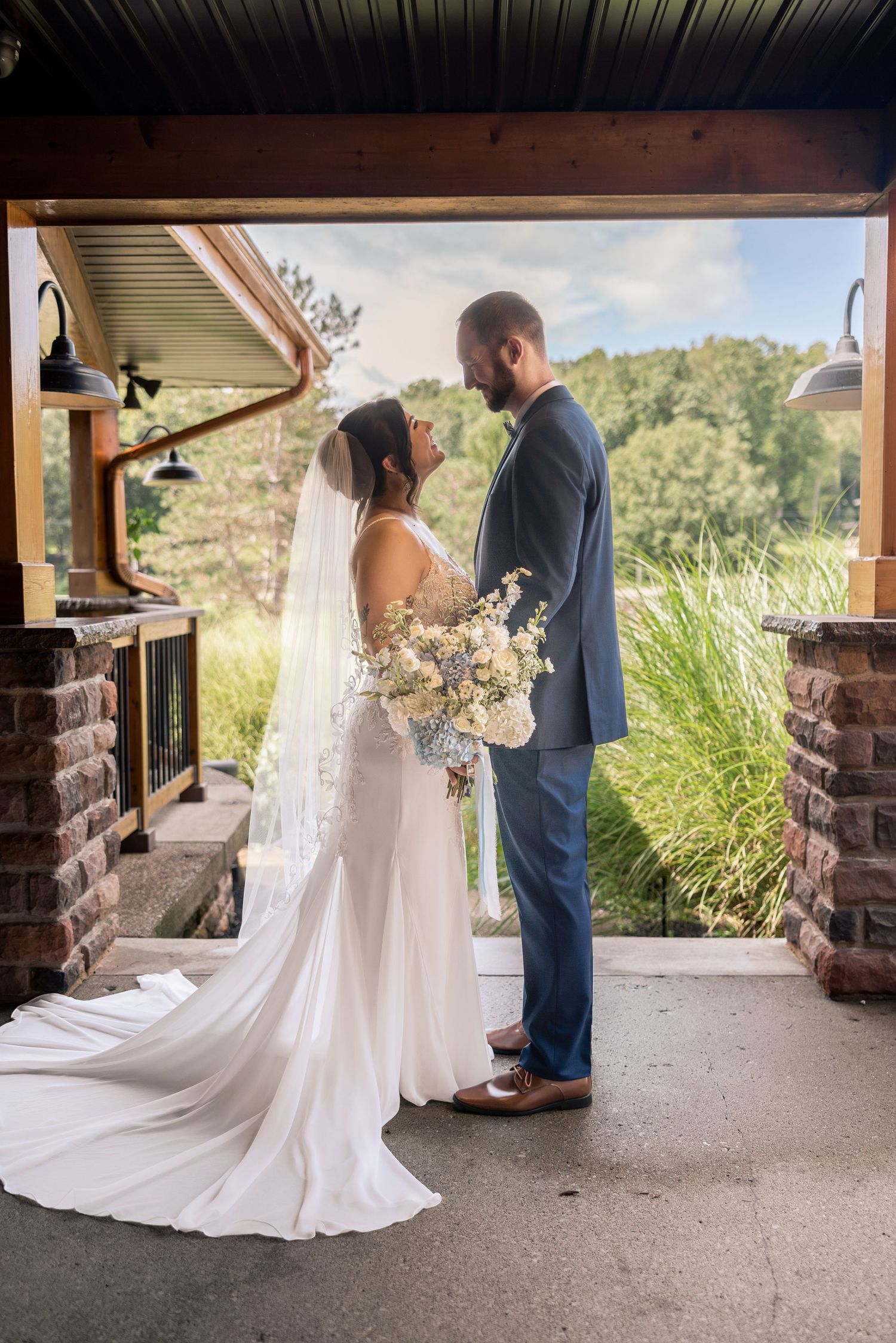 Bride in white gown with veil and groom in blue suit share an intimate moment under a rustic wooden pavilion outdoors.