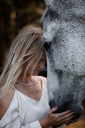 Blonde woman in white dress tenderly touching a dapple grey horse face in an intimate equine portrait.