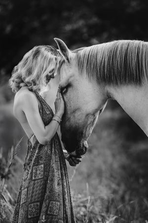 Black and white portrait of a woman in floral dress sharing a moment with a white horse.