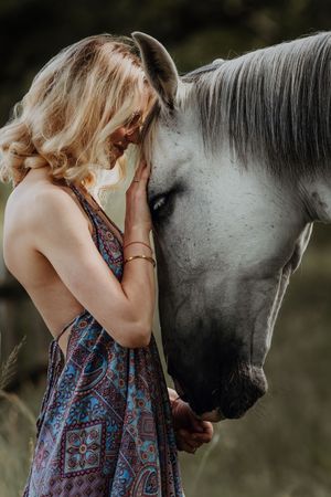 Blonde woman in bohemian dress tenderly embracing a dapple grey horse outdoors.