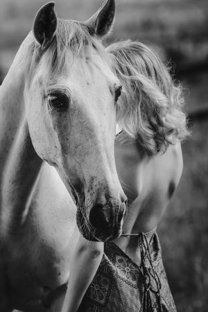 Black and white photo of a woman with curly blonde hair standing beside a white horse in a field.