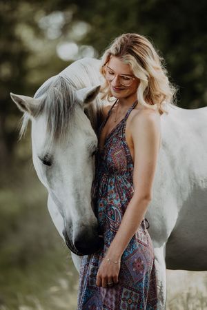 Blonde woman in boho dress standing beside a white horse outdoors.