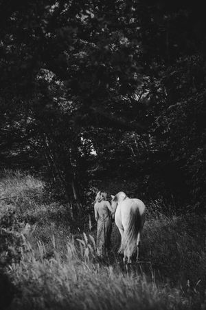 Black and white image of two people standing beside a white horse at the edge of a dark forest.