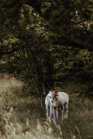 A woman in a flowing dress embraces a white horse in a lush green woodland clearing.
