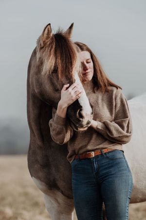 Young woman in brown sweater tenderly embracing a gray horse outdoors.