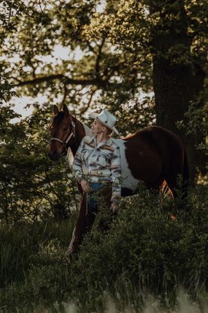 A blonde woman in a cowboy hat and western shirt leads a chestnut pinto horse through lush green woods.