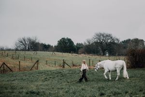 A person leads a white horse across a green pasture on an overcast day.