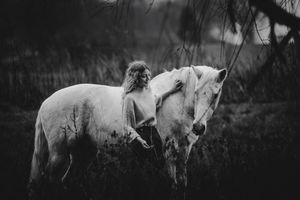 A black and white photo of a woman gently embracing a white horse among wild shrubs and branches.