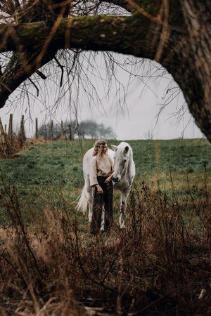 Woman in cream sweater standing with a white horse in a green field framed by bare tree branches.