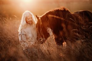 Young girl with long blonde hair gently touching a dark horse in a golden sunlit field.