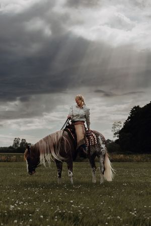 Woman riding a spotted horse in a green field under dramatic stormy skies.