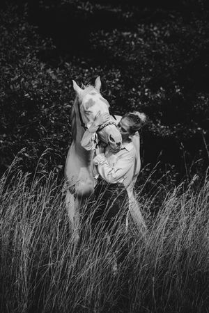 Black and white photo of a woman kissing a white horse in a field of tall grass with dark foliage background.