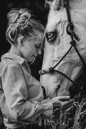 Black and white photo of a woman tenderly bonding with a white horse, holding its lead rope in a serene outdoor setting.