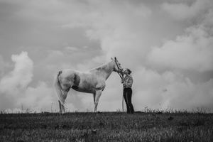 Black and white photo of a person standing with a white horse on a grassy hill under a cloudy sky.
