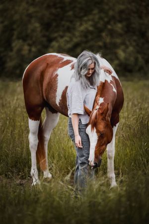 Woman with gray hair bonding with a brown and white paint horse in a lush green field.