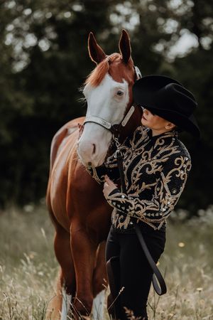 A woman in an ornate western show jacket and black hat stands closely with a chestnut horse in a meadow.