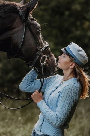 Woman in blue sweater and cap stands close to a dark bay horse while holding its bridle outdoors.