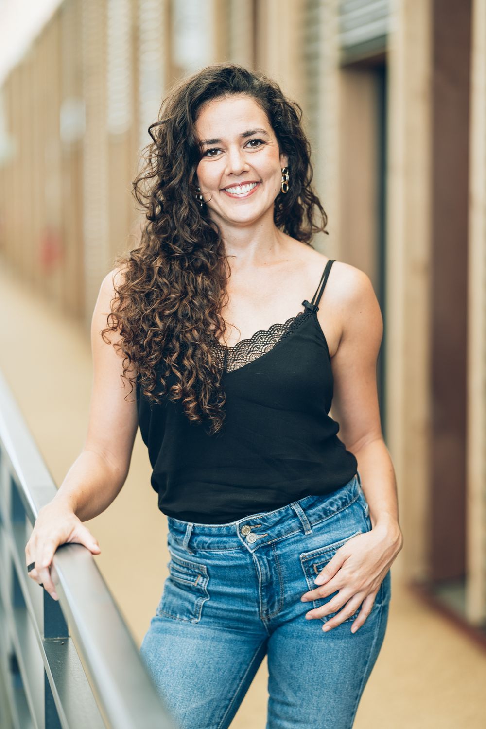 Smiling woman with curly brown hair in black cami top and jeans leaning on railing outdoors.