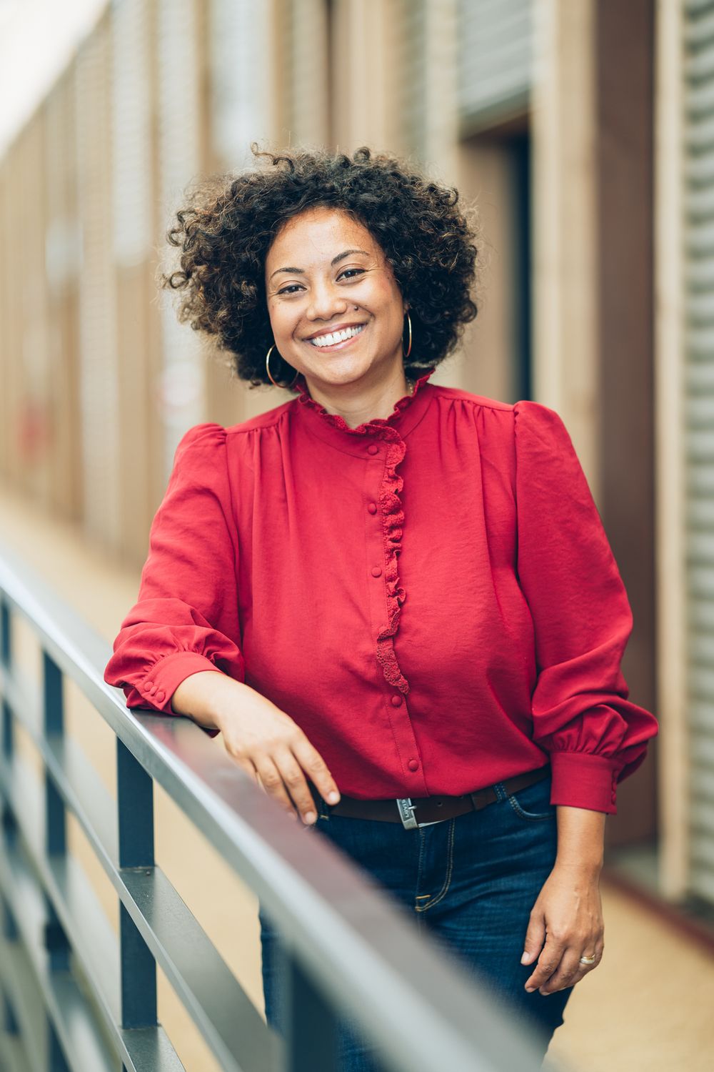Smiling woman with curly hair in red ruffled blouse leaning on railing in modern building corridor.