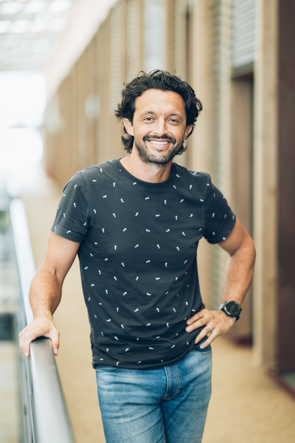 Smiling man with dark hair wearing a black patterned t-shirt and jeans, leaning on a railing outdoors.