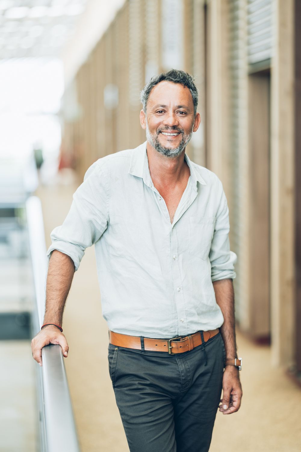 Middle-aged man with gray beard smiling confidently, wearing light blue shirt and brown belt, leaning on railing outdoors.