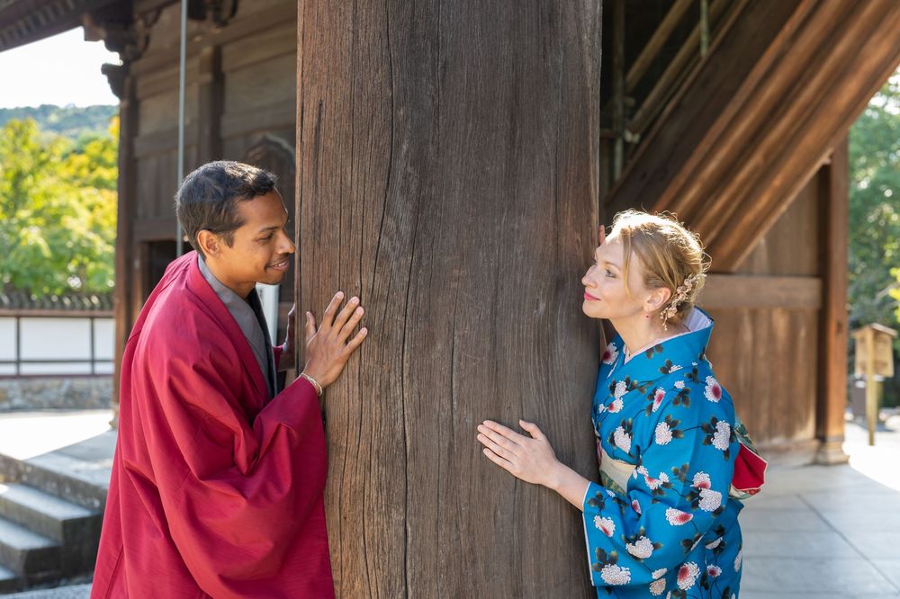 Two people in traditional Japanese kimonos converse beside a large wooden pillar at a historic temple.