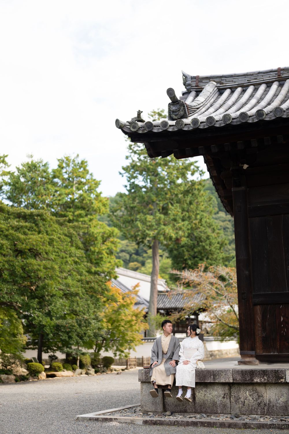 Couple in traditional Japanese kimono sitting beside an ancient temple gate surrounded by lush green trees in Kyoto.