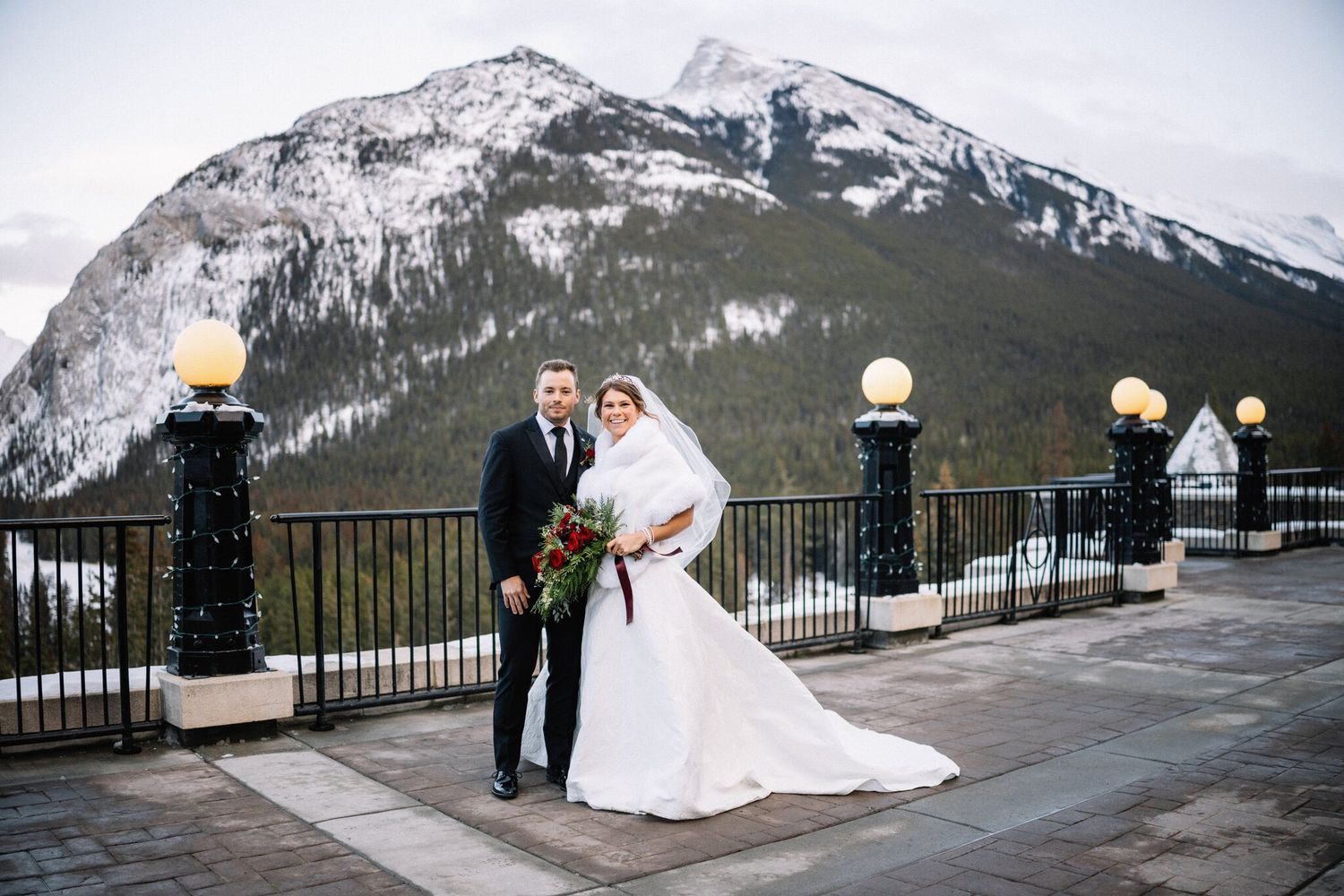 Smiling bride and groom with a red rose bouquet on a Fairmont Banff Springs terrace with mountains behind