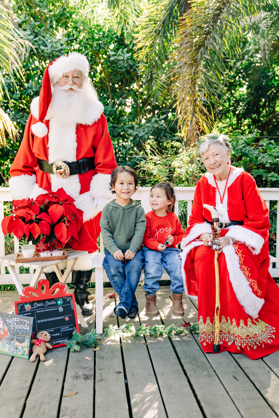 Santa and Mrs. Claus photos in Lake Worth Beach with two children surrounded by tropical greenery and Christmas decorations.