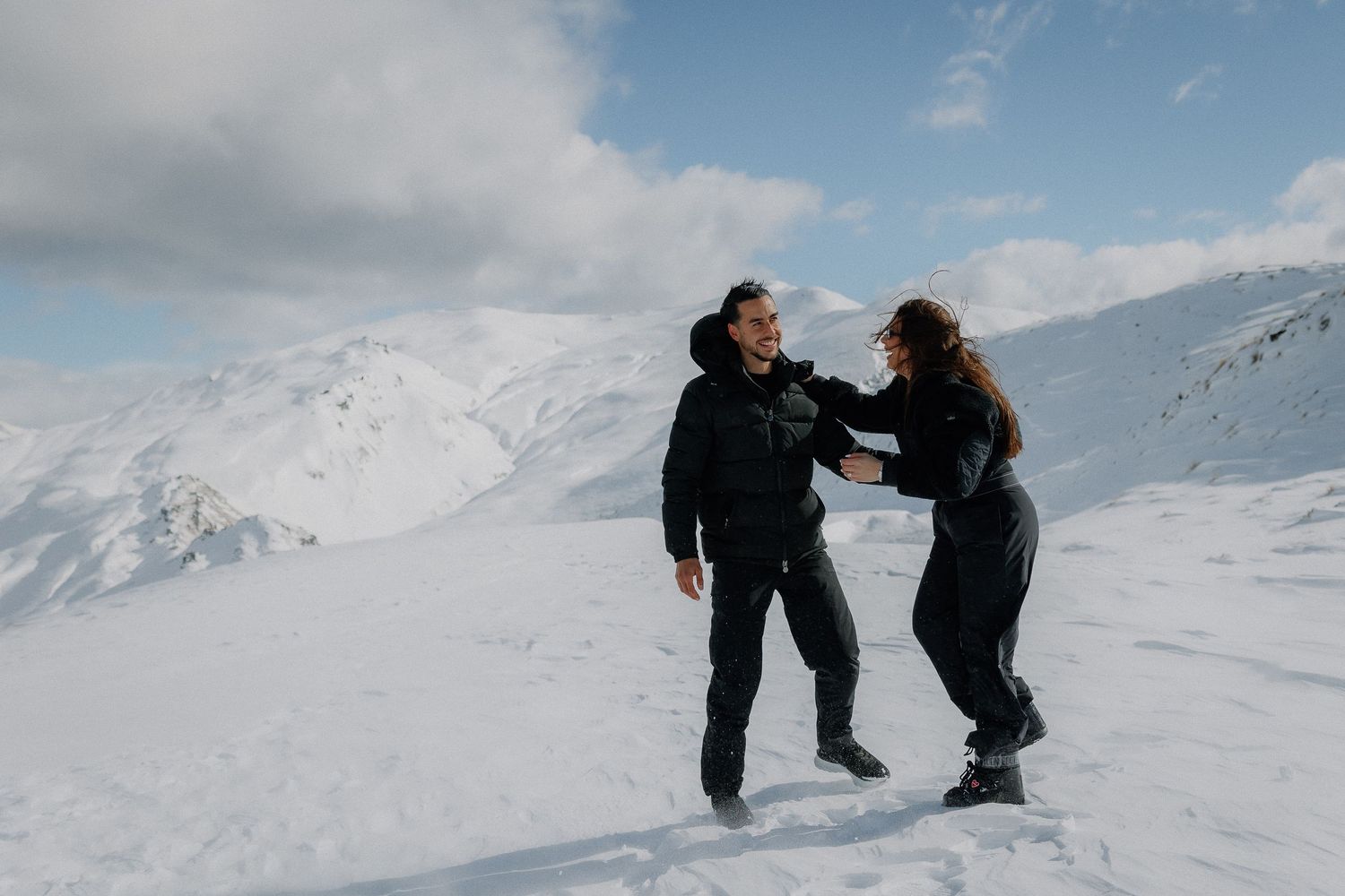 Couple laughing and playing in snow on a mountain peak, wearing black winter outfits with snowy peaks behind them.