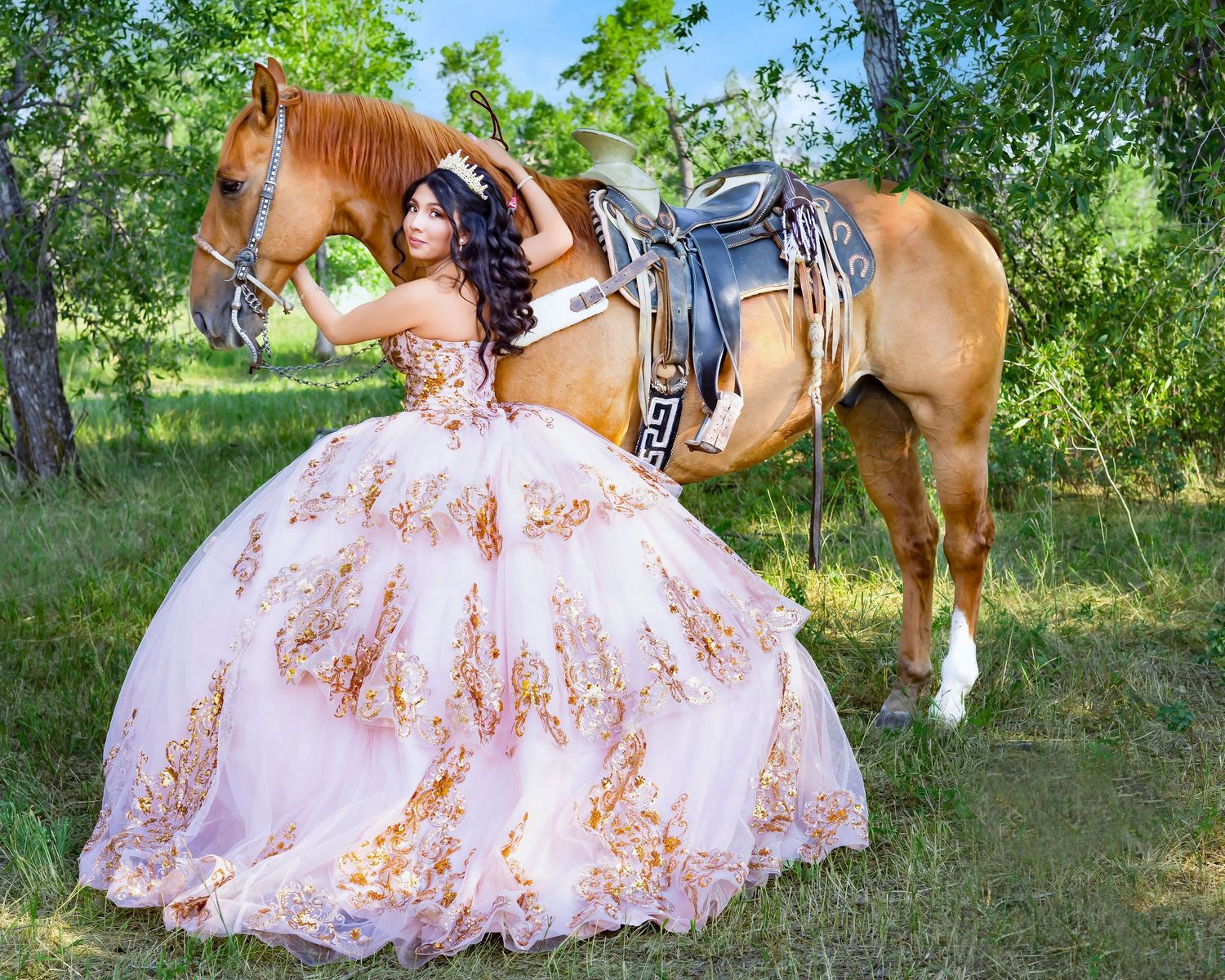 Young woman in pink and gold quinceañera ball gown poses beside a saddled brown horse in a lush green outdoor setting.