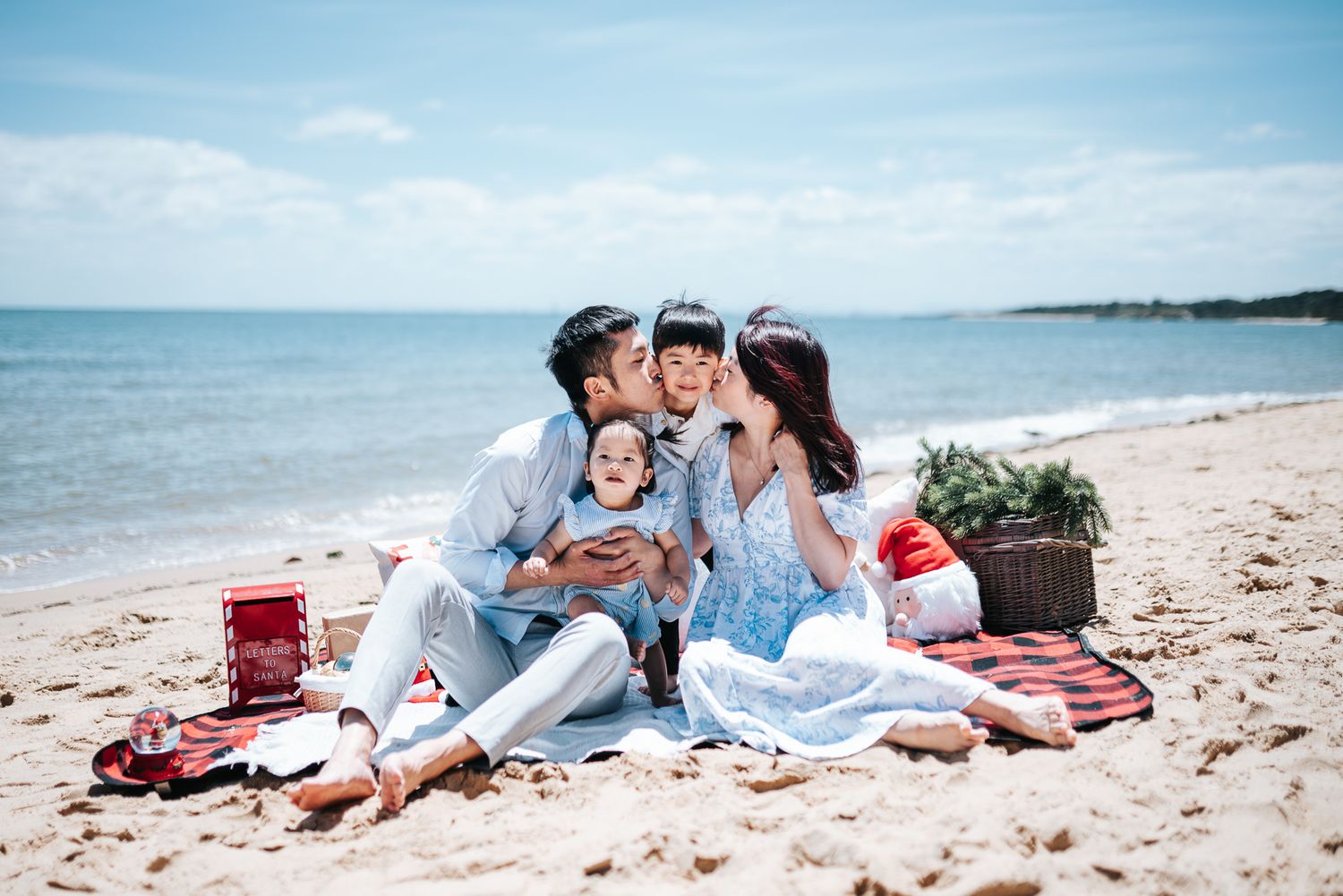 Family Christmas beach picnic portrait with parents kissing baby on cheeks taken during a Melbourne Christmas Mini Session by Pei A.