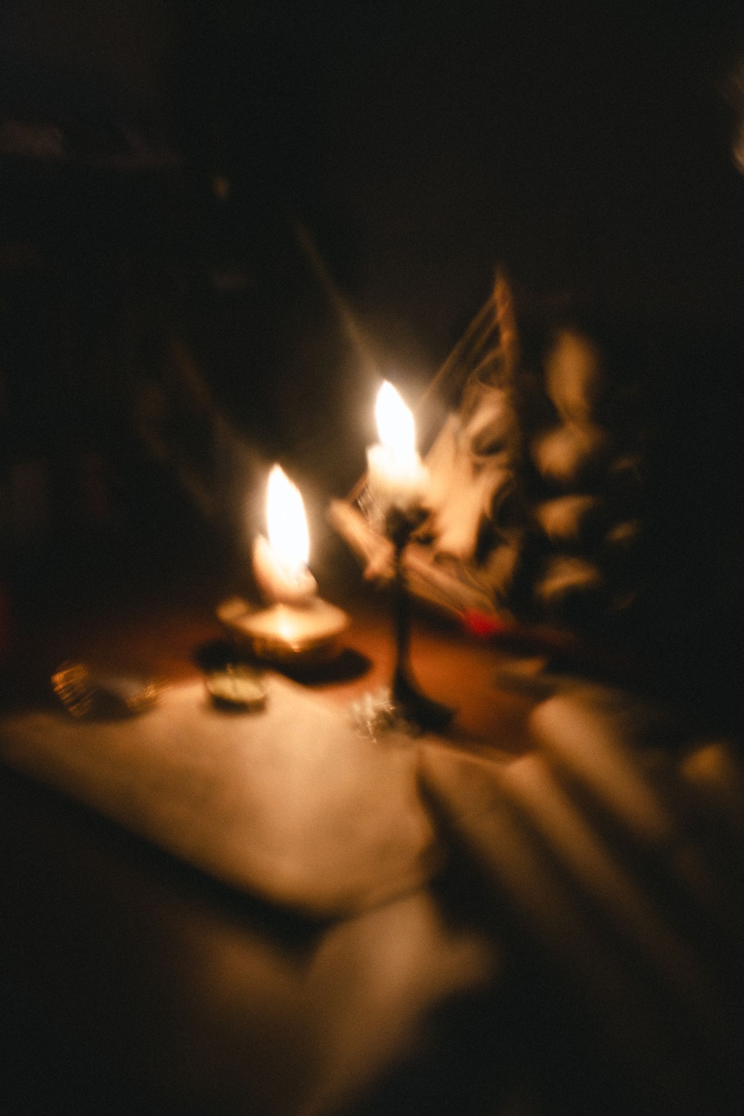Two glowing candles illuminate a dark table with small objects in a moody, atmospheric candlelit scene.