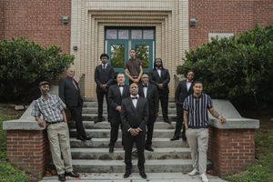 A group of well-dressed men in suits and formal attire pose on brick steps outside a building.