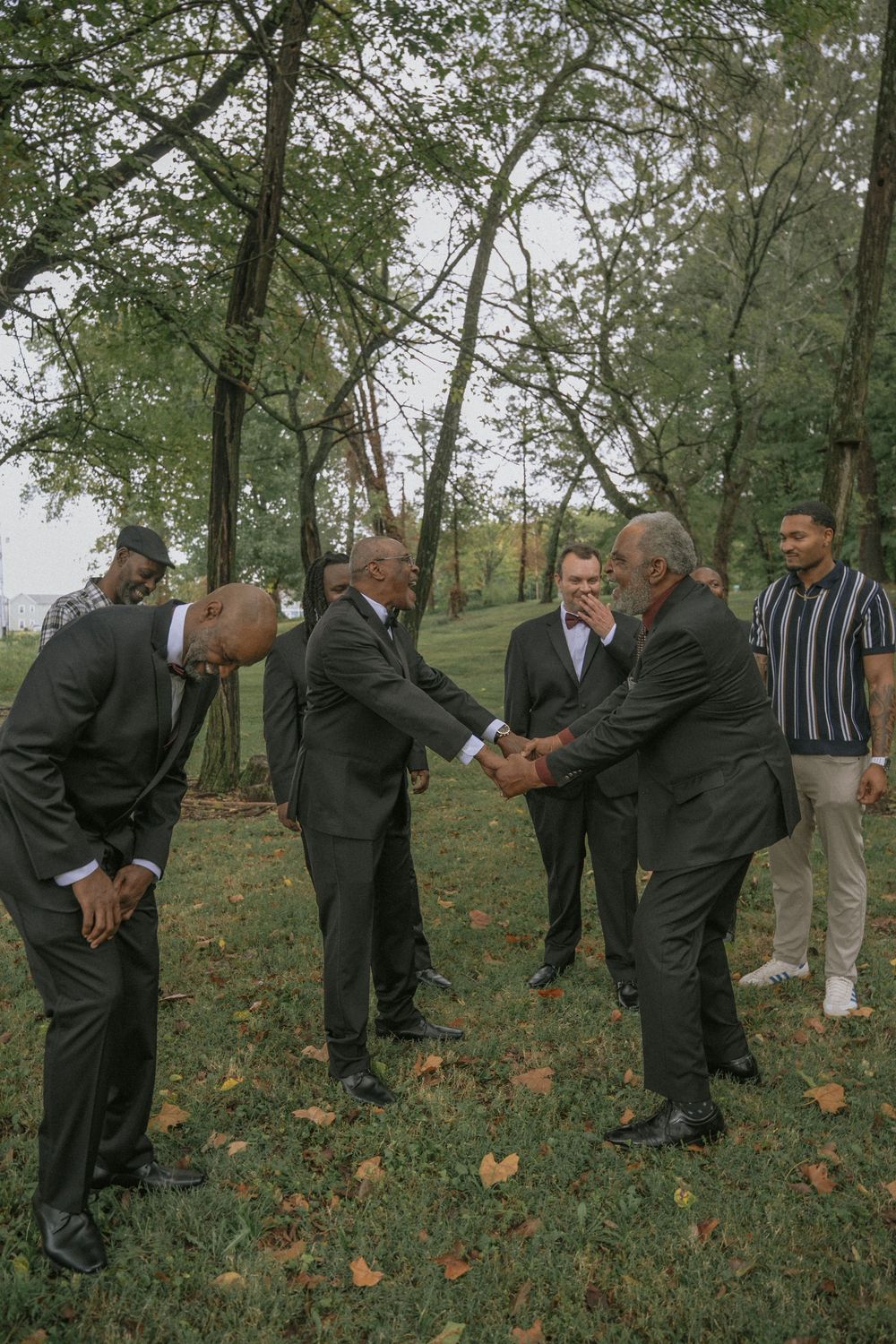 Group of men in dark suits shaking hands outdoors in a park setting with trees and fallen leaves.
