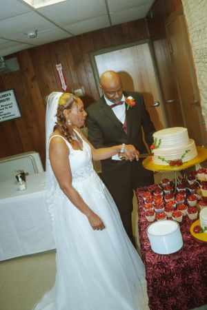 Bride and groom cutting their white wedding cake together at their reception, surrounded by elegant red and white cupcakes.