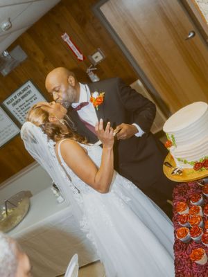 Bride and groom sharing a kiss while cutting their white wedding cake decorated with orange roses.