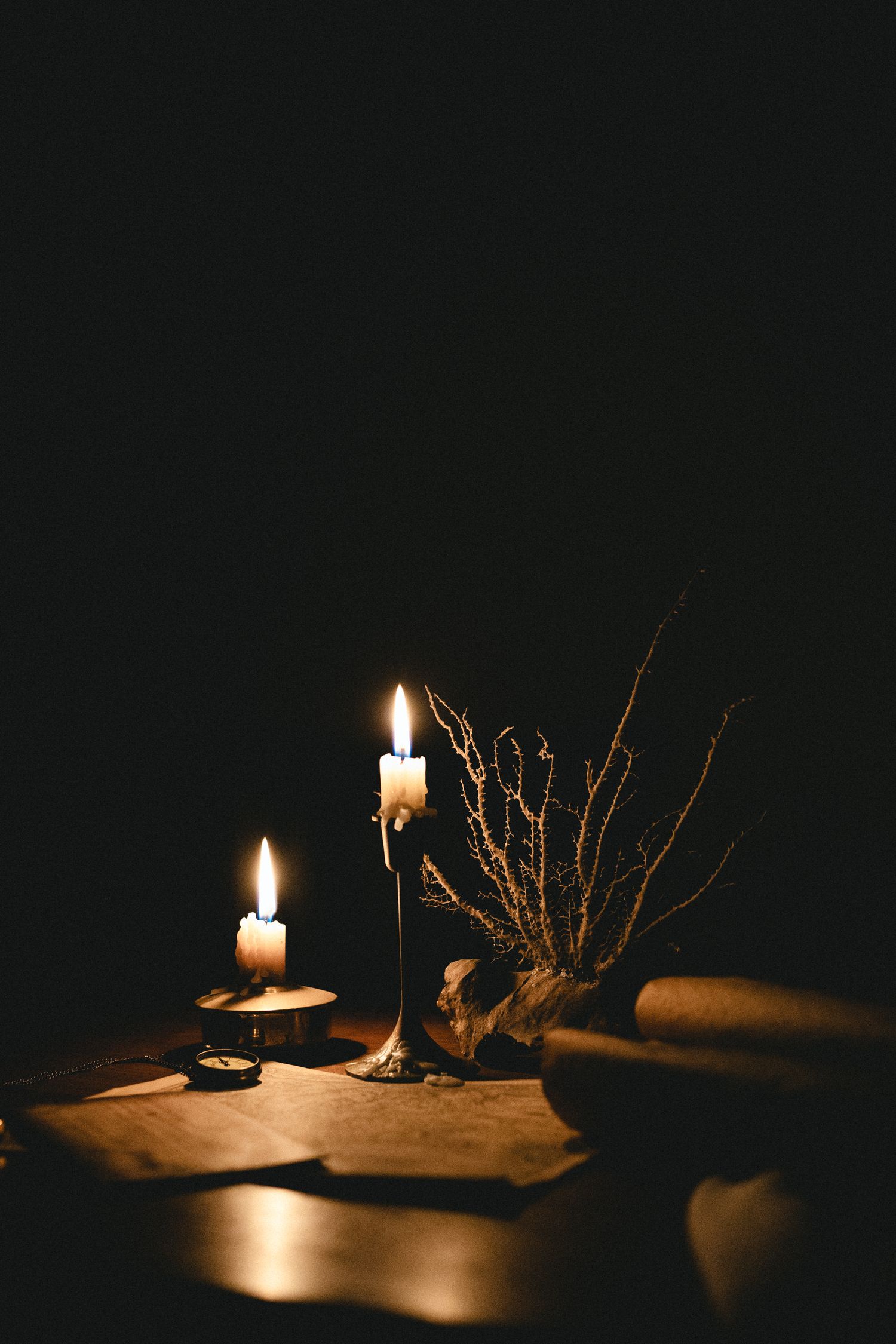 Two lit candles on a dark table with dried branches and aged parchment paper in moody candlelight.