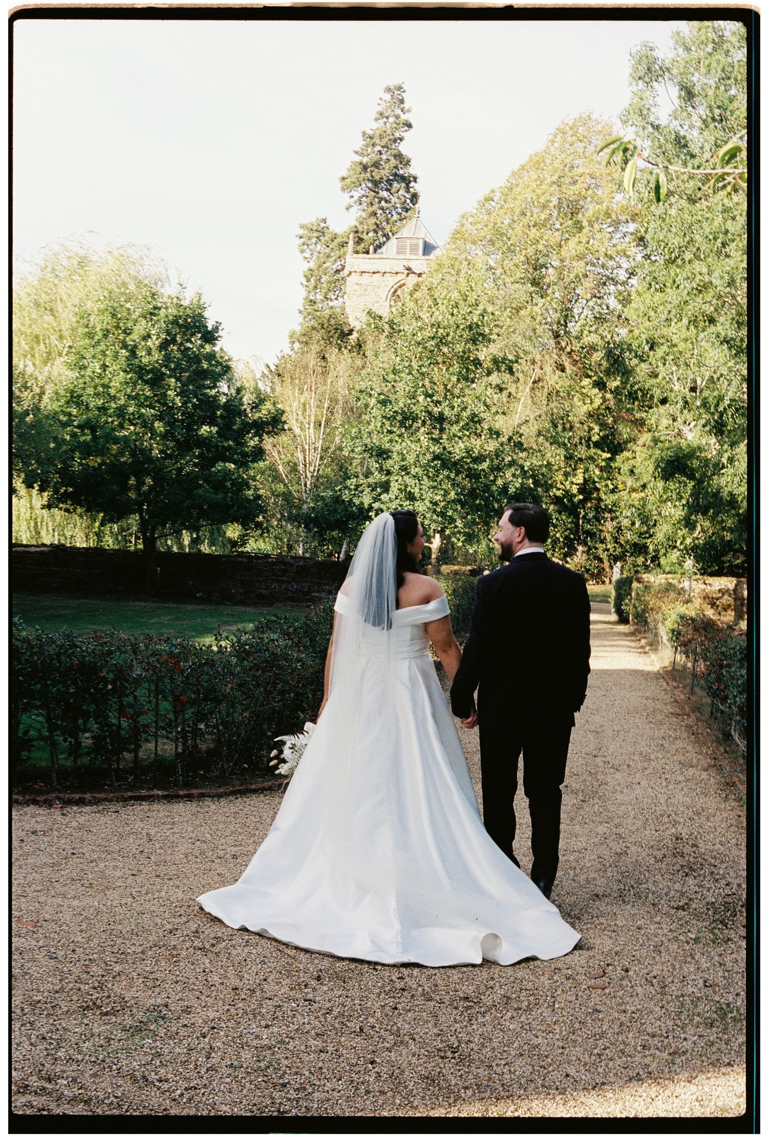Bride and groom walking hand in hand along a gravel garden path, bride in white gown with veil and train.