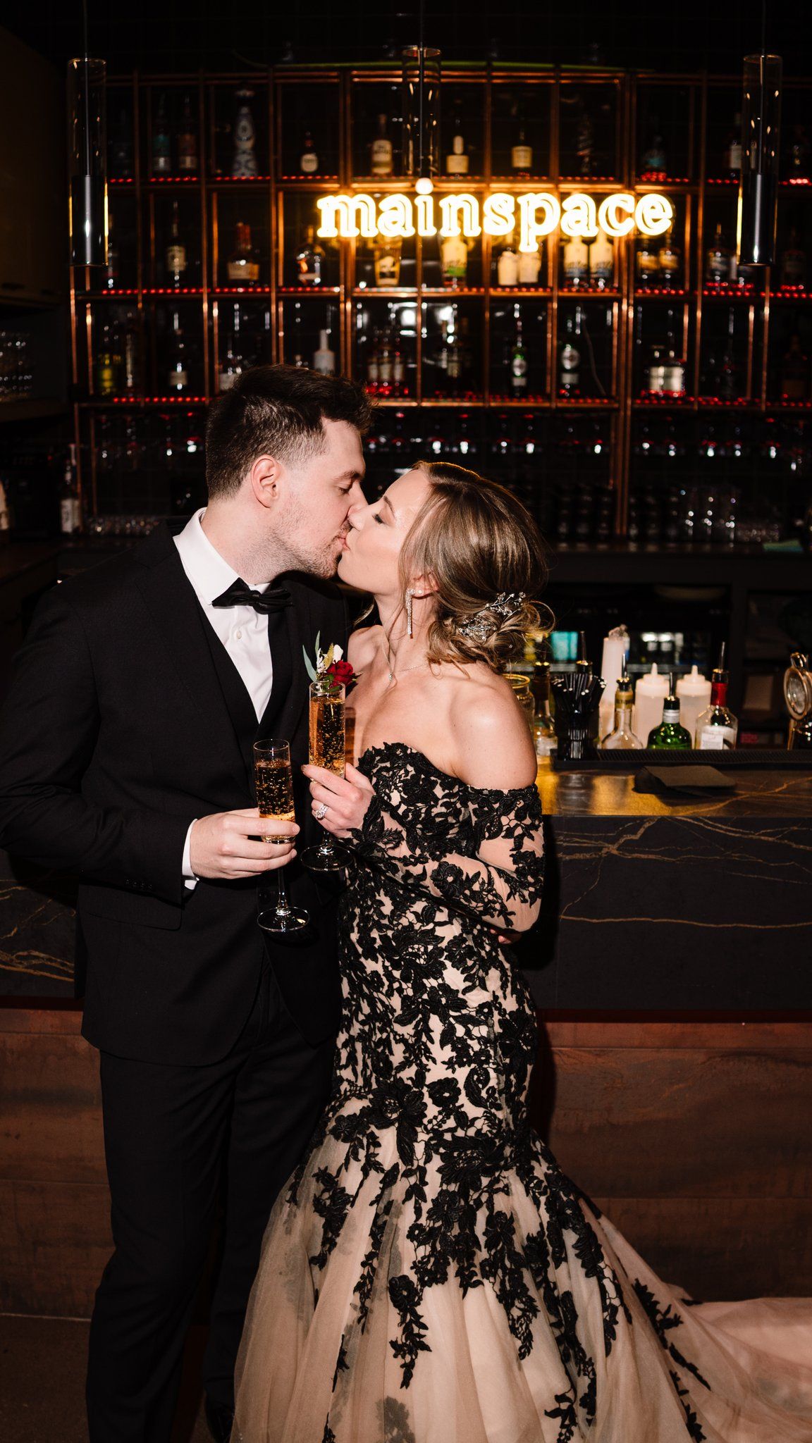 Couple kissing with champagne glasses under the glowing Mainspace neon sign at the bar