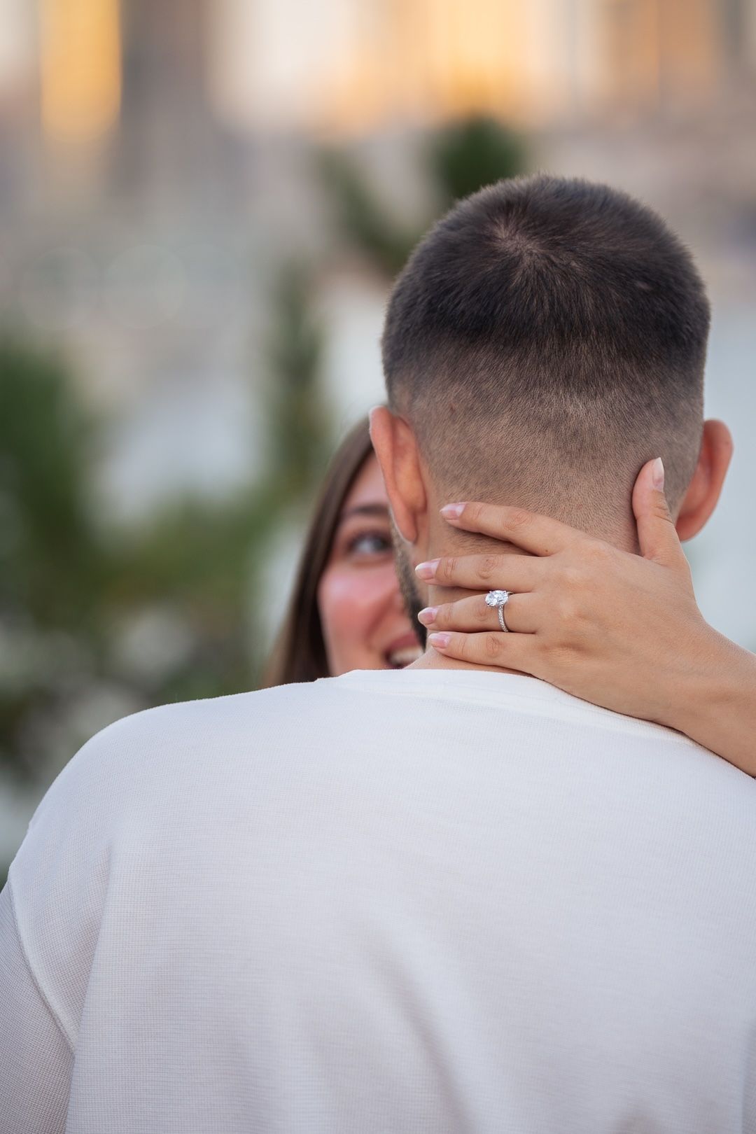A woman smiling and embracing a man from behind, showing off her diamond engagement ring on his neck.