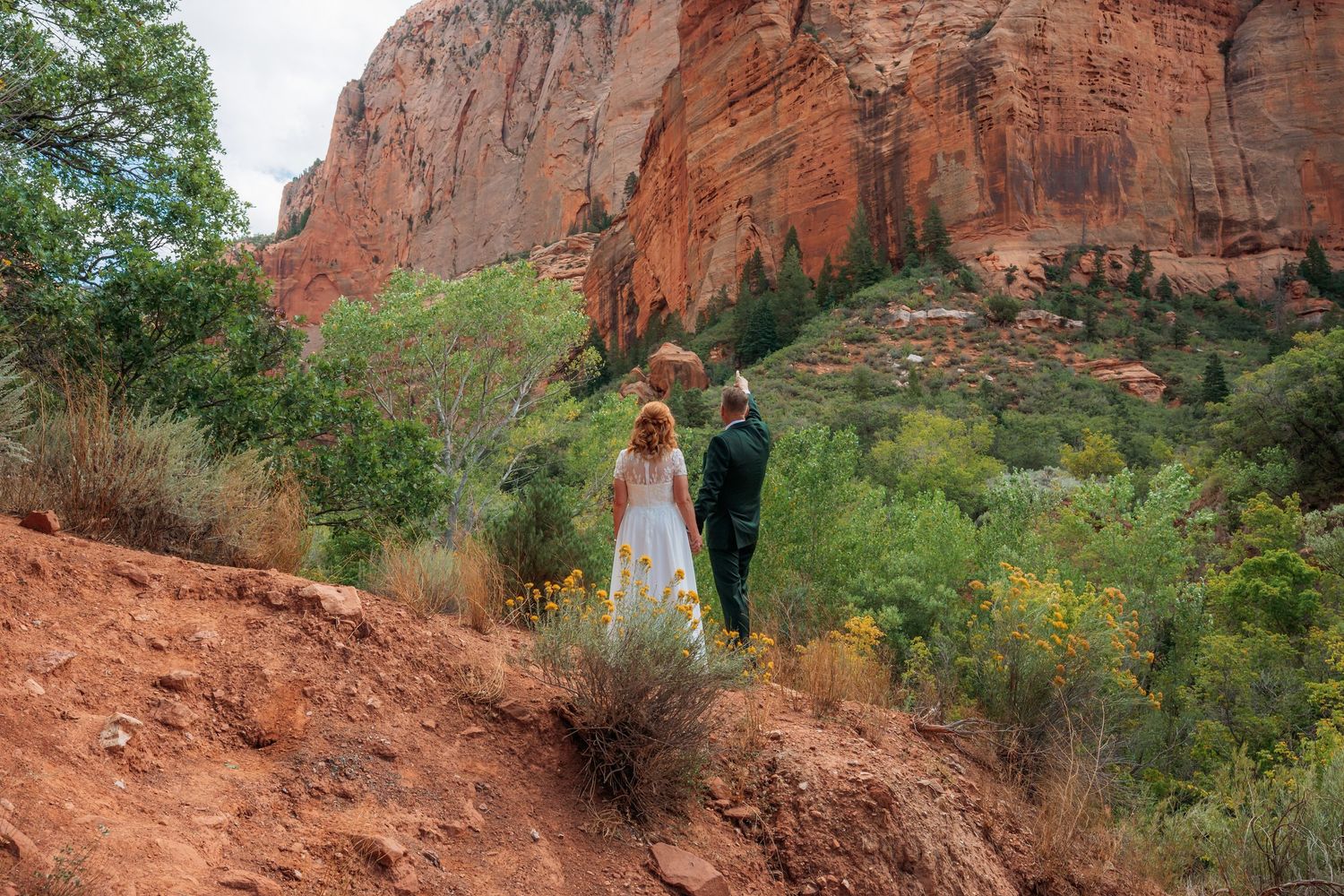 A bride and groom stand on a red rock trail in Sedona Arizona surrounded by lush greenery and towering sandstone cliffs.