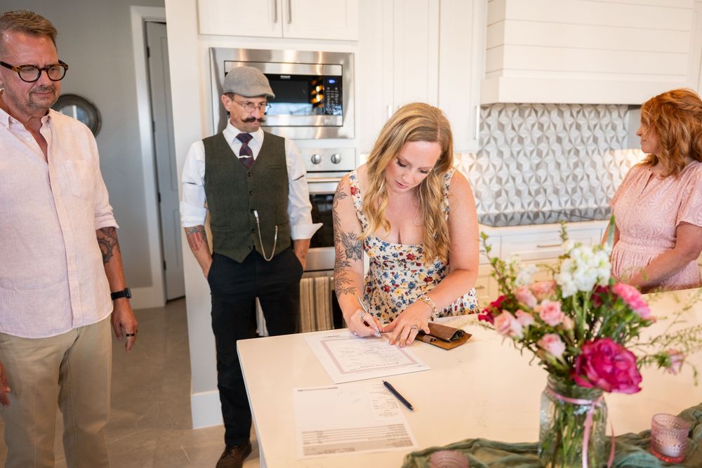 Blonde woman signing documents at a kitchen table with three people watching during a home signing ceremony.