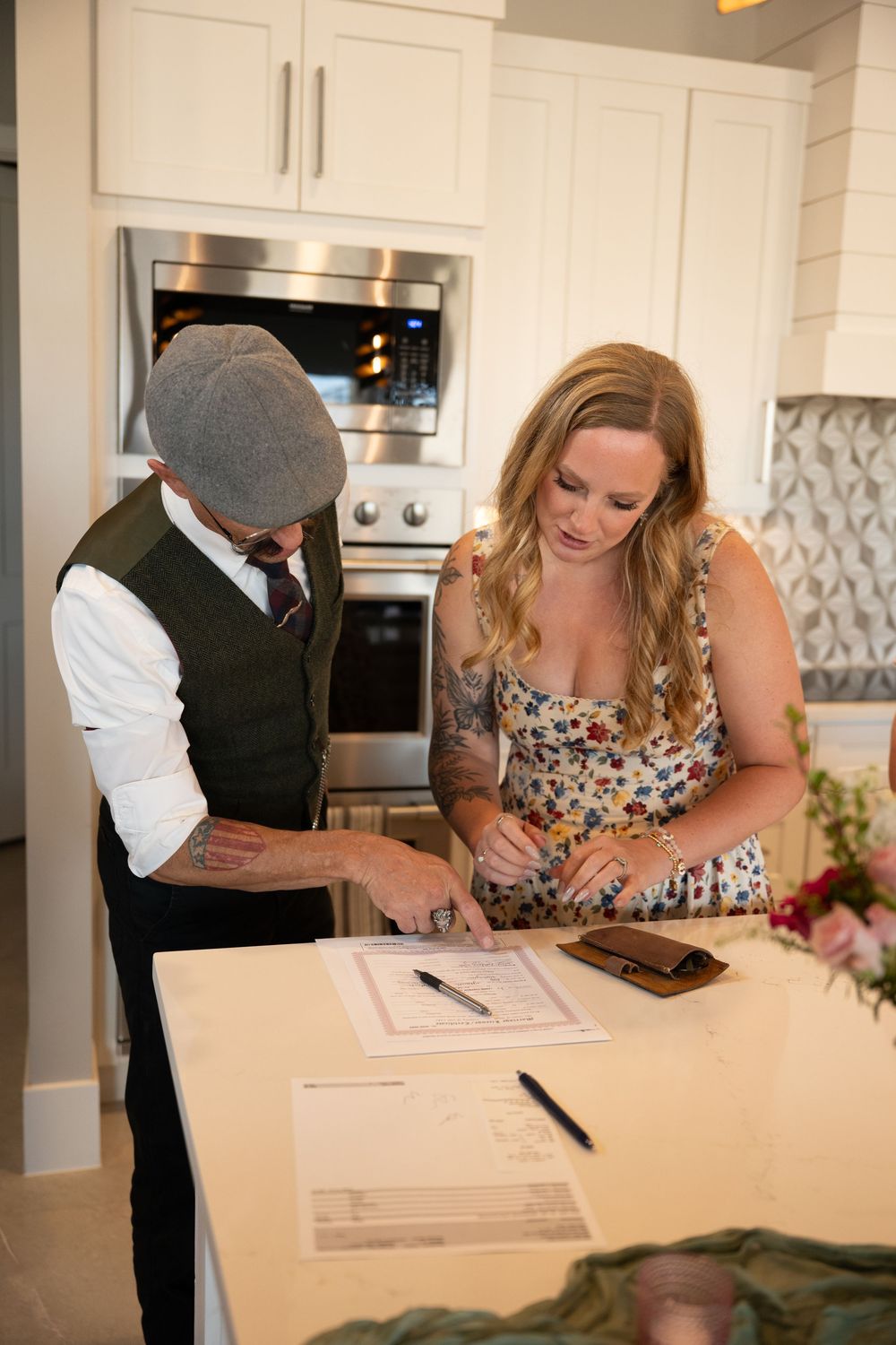 A man and woman review and sign documents together at a kitchen counter in a modern white kitchen.