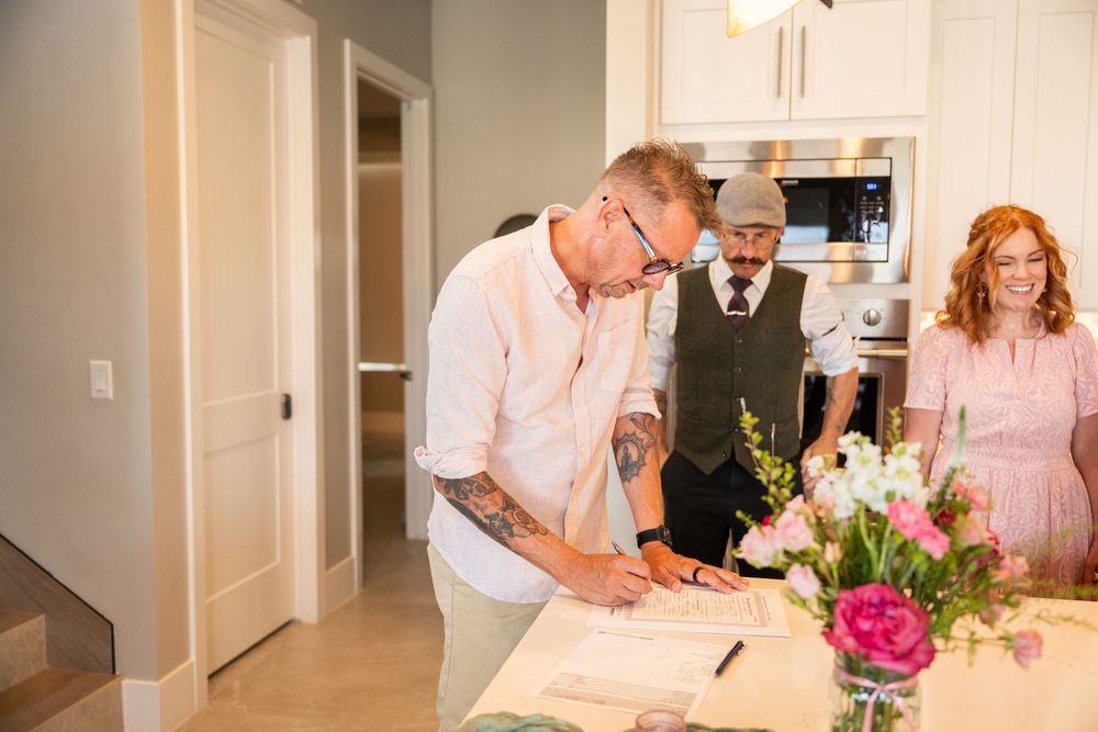 Man signing documents at a kitchen counter during a home closing ceremony with two witnesses standing nearby.