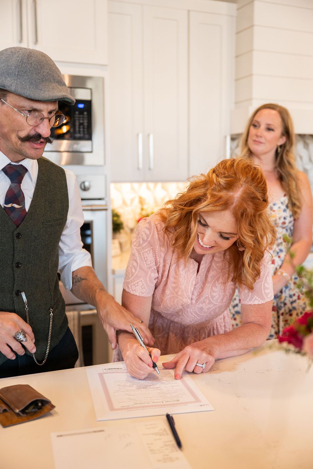 Bride with red hair signs marriage license in modern kitchen with officiant in vintage vest and flat cap.