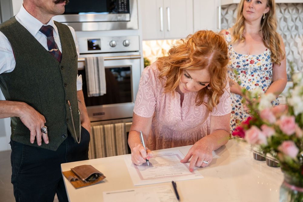 A redhead woman signs documents at a kitchen counter while a man in a vest and woman in floral dress watch.